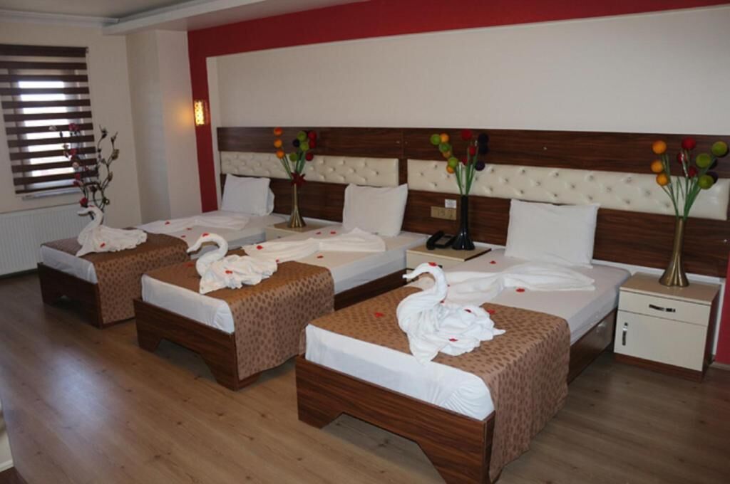 A hotel room with three beds, each with a swan-shaped towel and rose petals. There are bedside tables with lamps and a small cabinet.