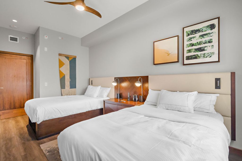 Une chambre d’hôtel avec deux lits, une armoire en bois, un ventilateur de plafond et deux œuvres d’art encadrées accrochées au mur.