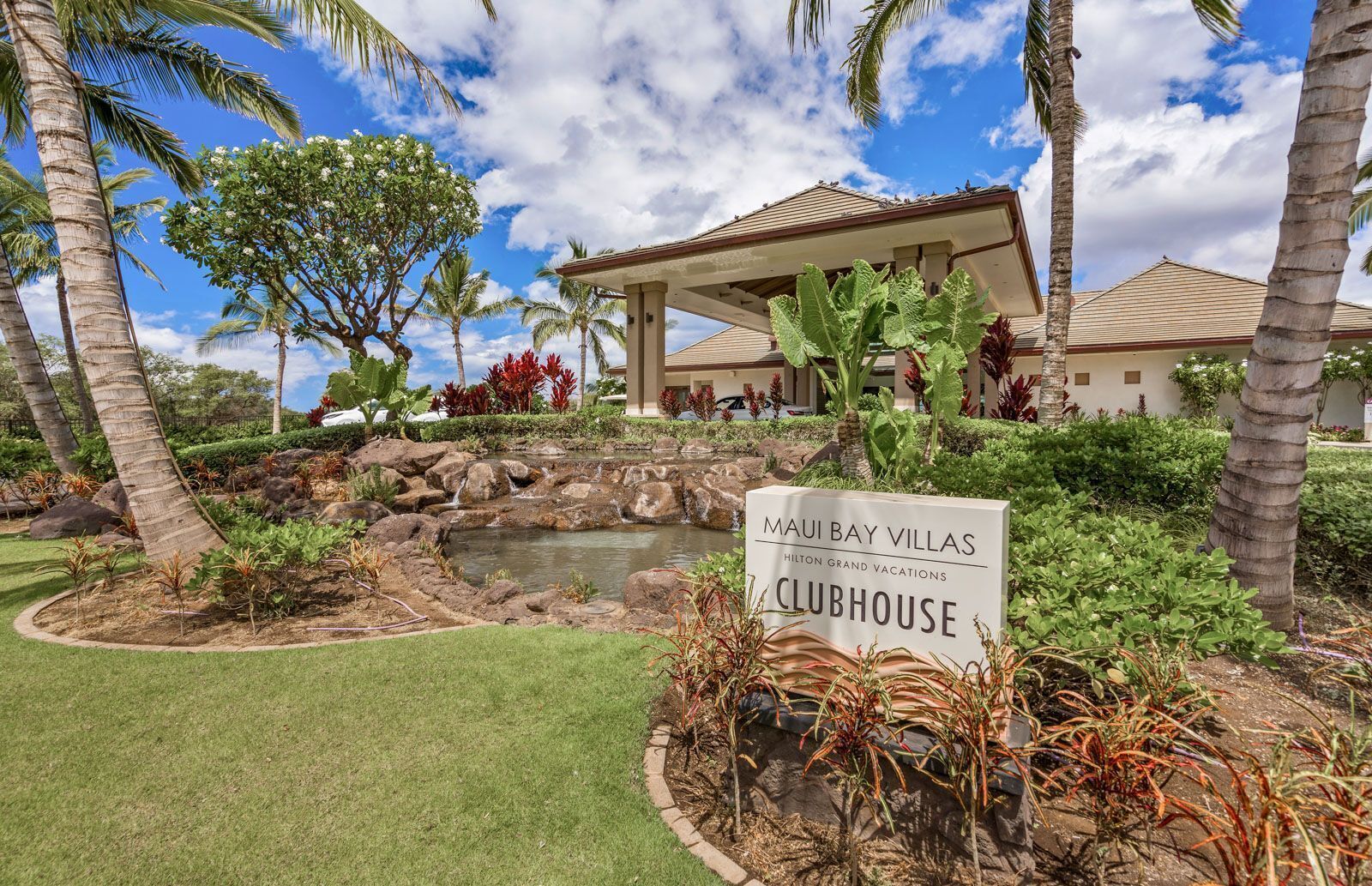 A sign for 'Maui Bay Villas Clubhouse' with a landscaped garden featuring palm trees, a small pond, and a gazebo.