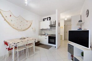 A compact kitchen with white cabinets, a dining table with chairs, a TV, and a net decoration on the wall. - Apartment 'Casa Perla Blu' with Mountain View, Wi-Fi and Air Conditioning (San Vito Lo Capo)