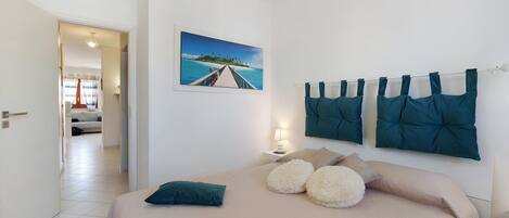 A bedroom with a bed, pillows, a nightstand, a lamp, a painting, and a view of a hallway.