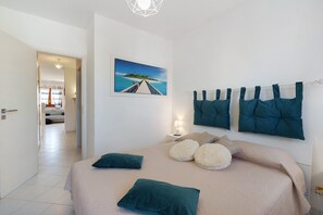 A bedroom with a bed, pillows, a nightstand, a lamp, a painting, and a view of a hallway. - Apartment 'Casa Perla Blu' with Mountain View, Wi-Fi and Air Conditioning (San Vito Lo Capo)