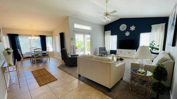 A spacious living area with a sofa, a coffee table, and a TV. There is a dining area with a table and chairs. The room has a ceiling fan and large windows. - Comfy-cozy 1BR home in gated 55+ community (Goodyear)