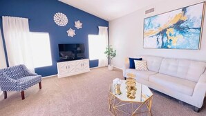 A living room with a TV, white sofa, patterned chair, decorative wall art, and a geometric coffee table. - Comfy-cozy 1BR home in gated 55+ community (Goodyear)