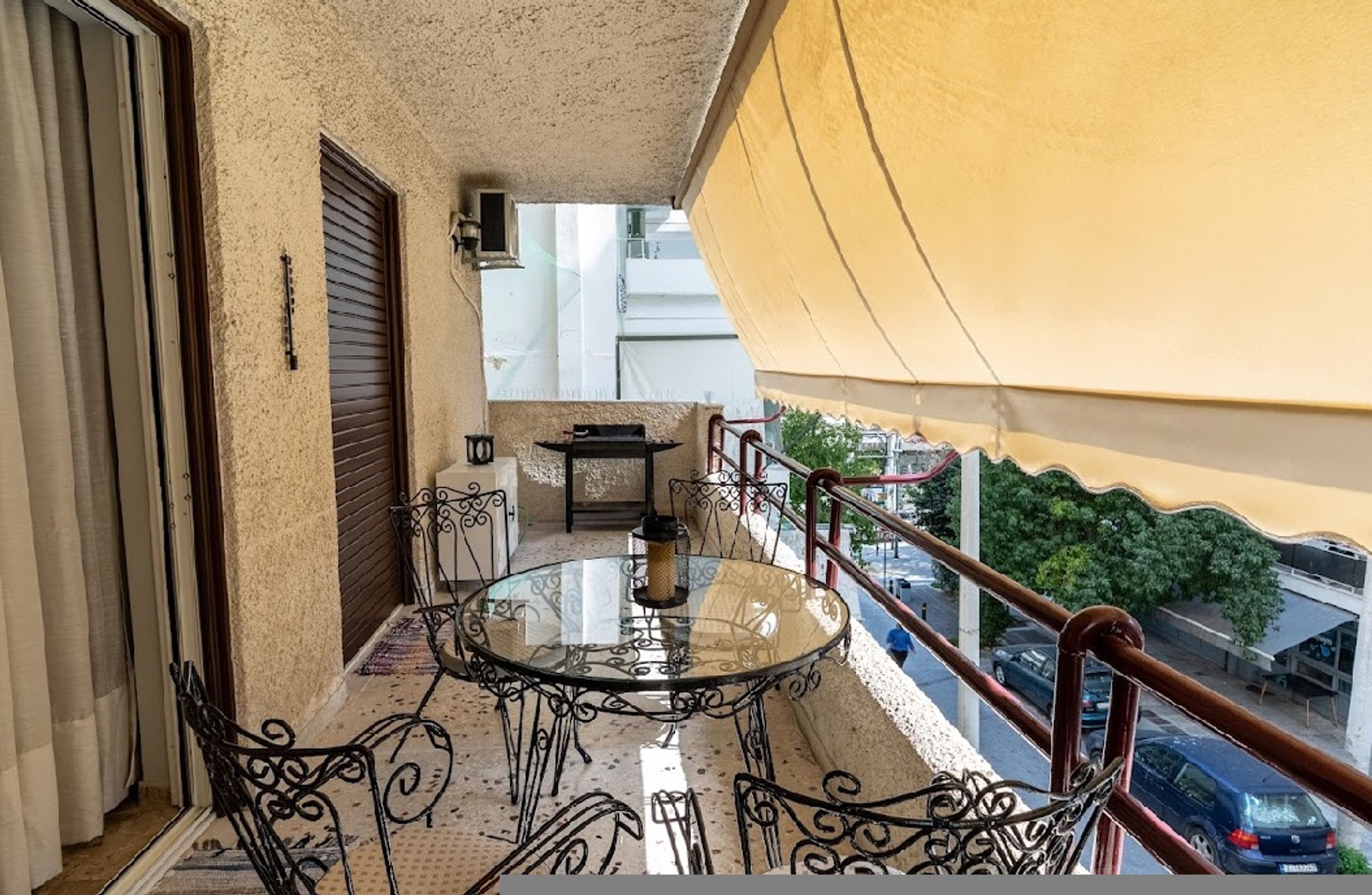 A balcony with a glass table and chairs, a view of the street, and a yellow awning.