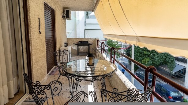 A balcony with a glass table and chairs, a view of the street, and a yellow awning. - Cozy Apartment in Faliro (Paleo Faliro)