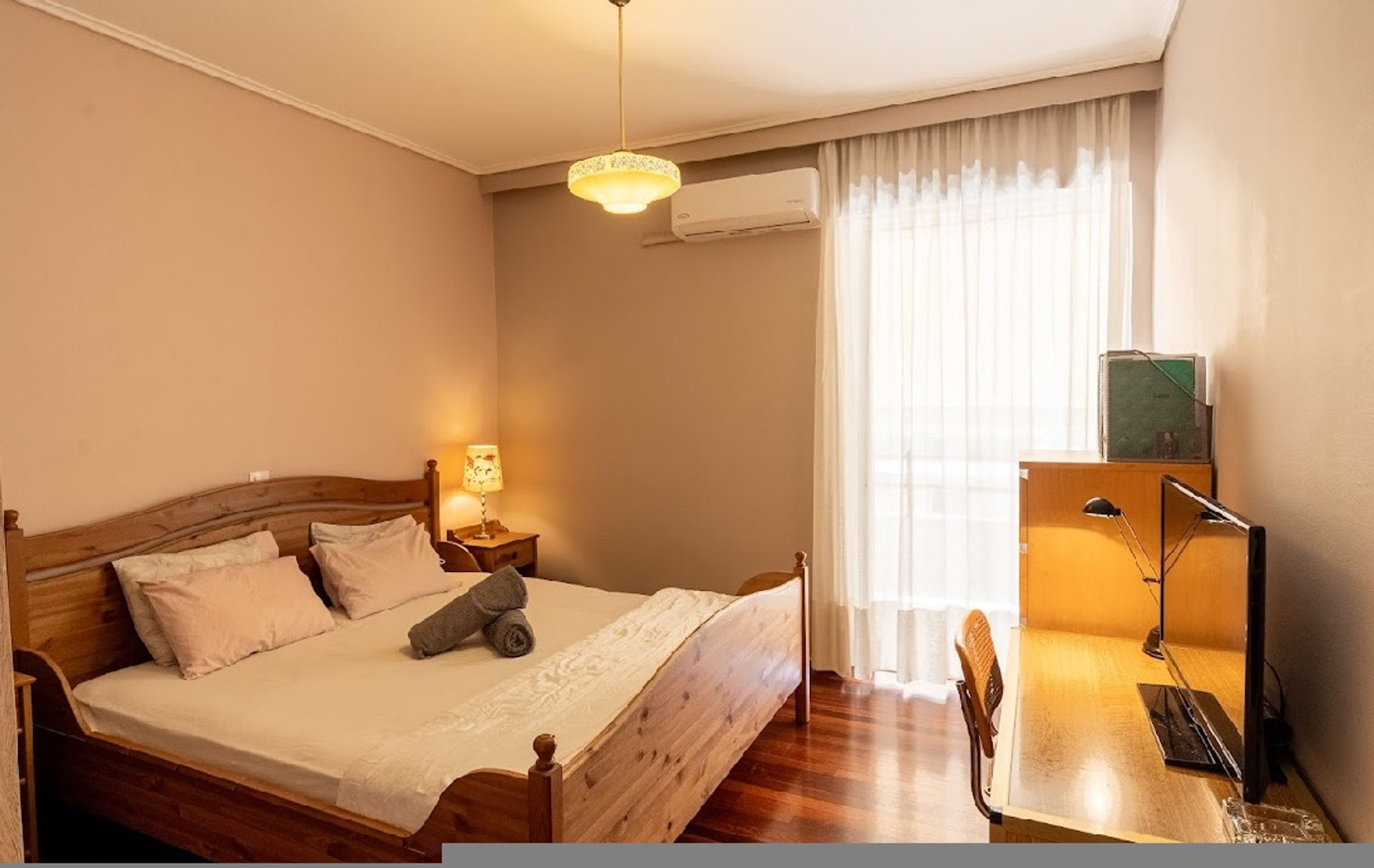 A bedroom with a wooden bed, a desk with a chair, a lamp, and a window with curtains.