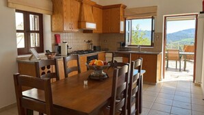 A kitchen with wooden cabinets, a dining table with chairs, a fruit bowl, and a view of the outdoors. - St Visianos stone house (Filousa)