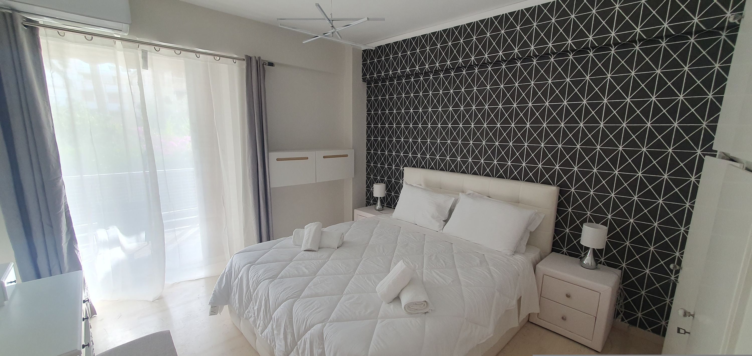 Een hotelkamer met een groot bed, nachtkastje, gordijnen voor het raam en een wand met een geometrisch patroon.