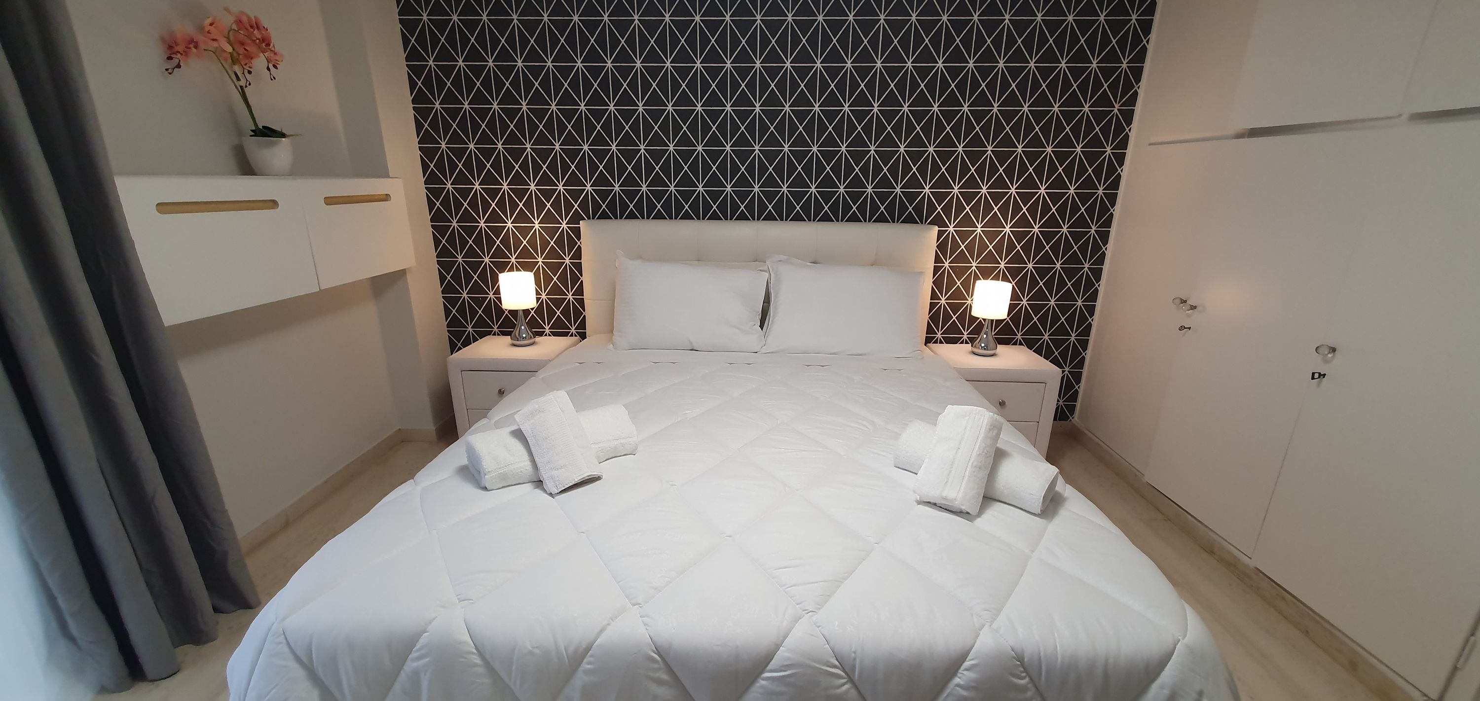 Een hotelkamer met een bed, twee nachtkastjes met lampen, een kledingkast en een muur met een geometrisch patroon.