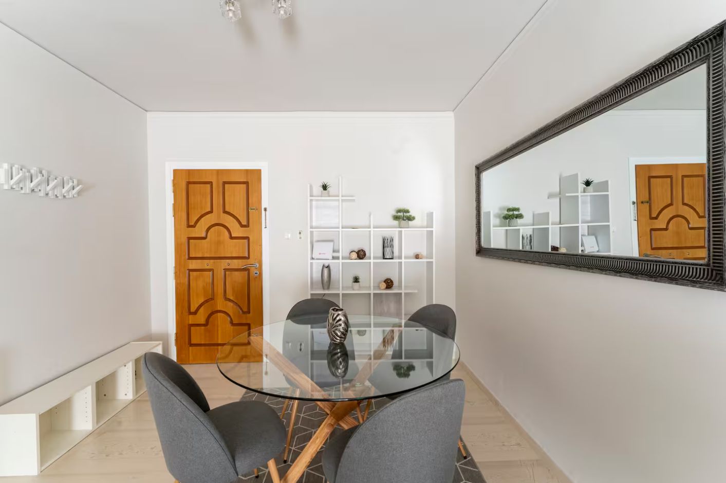 A modern dining area with a glass table and grey chairs, a wooden door, and a white shelving unit with decorative items.