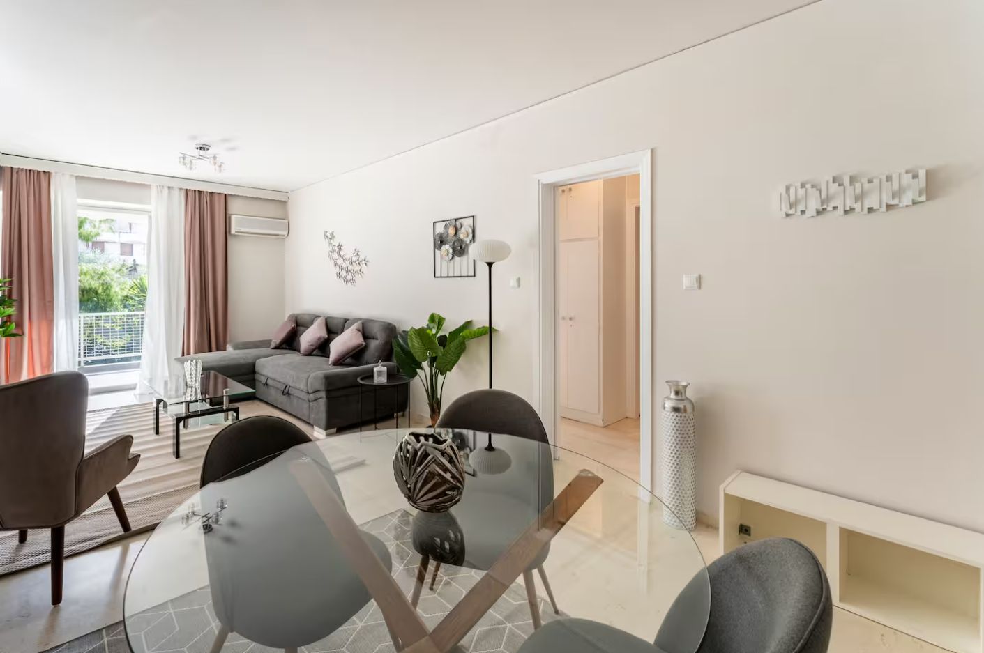 Een moderne woonkamer met een glazen eettafel, een grijze bank en een groot raam met gordijnen.