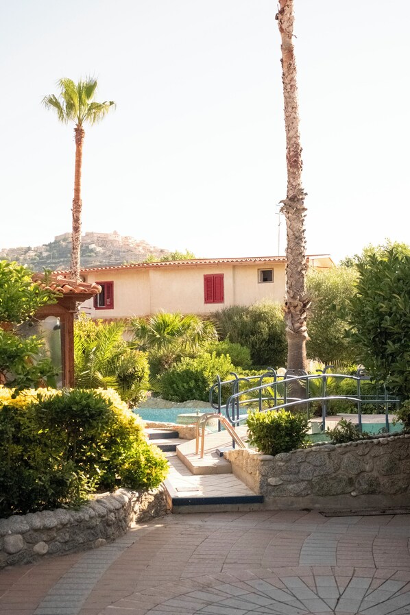 A pool area with steps leading to a pool, surrounded by palm trees and a building with red shutters.