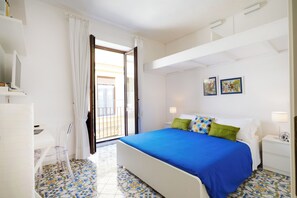 A bedroom with a bed, a desk, a chair, a nightstand, a lamp, and a window with curtains. - The Casket (Sorrento)