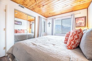 Ein Schlafzimmer mit Holzboden, einem Bett mit gemusterten Kissen, einem Kreuz an der Wand und einem Fenster mit Jalousien.