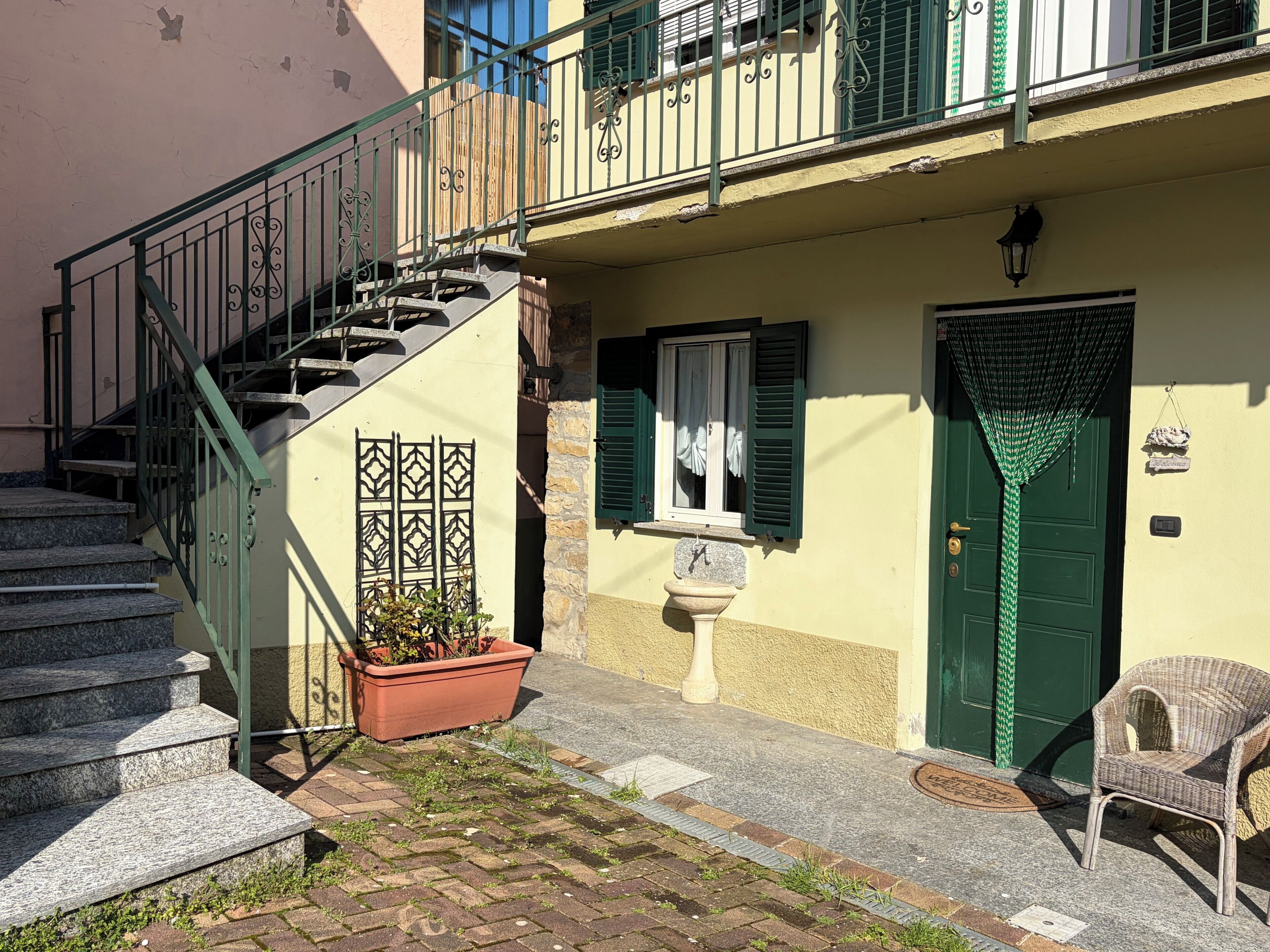 Un edificio con una escalera, una puerta verde, una planta en maceta y una silla de mimbre.