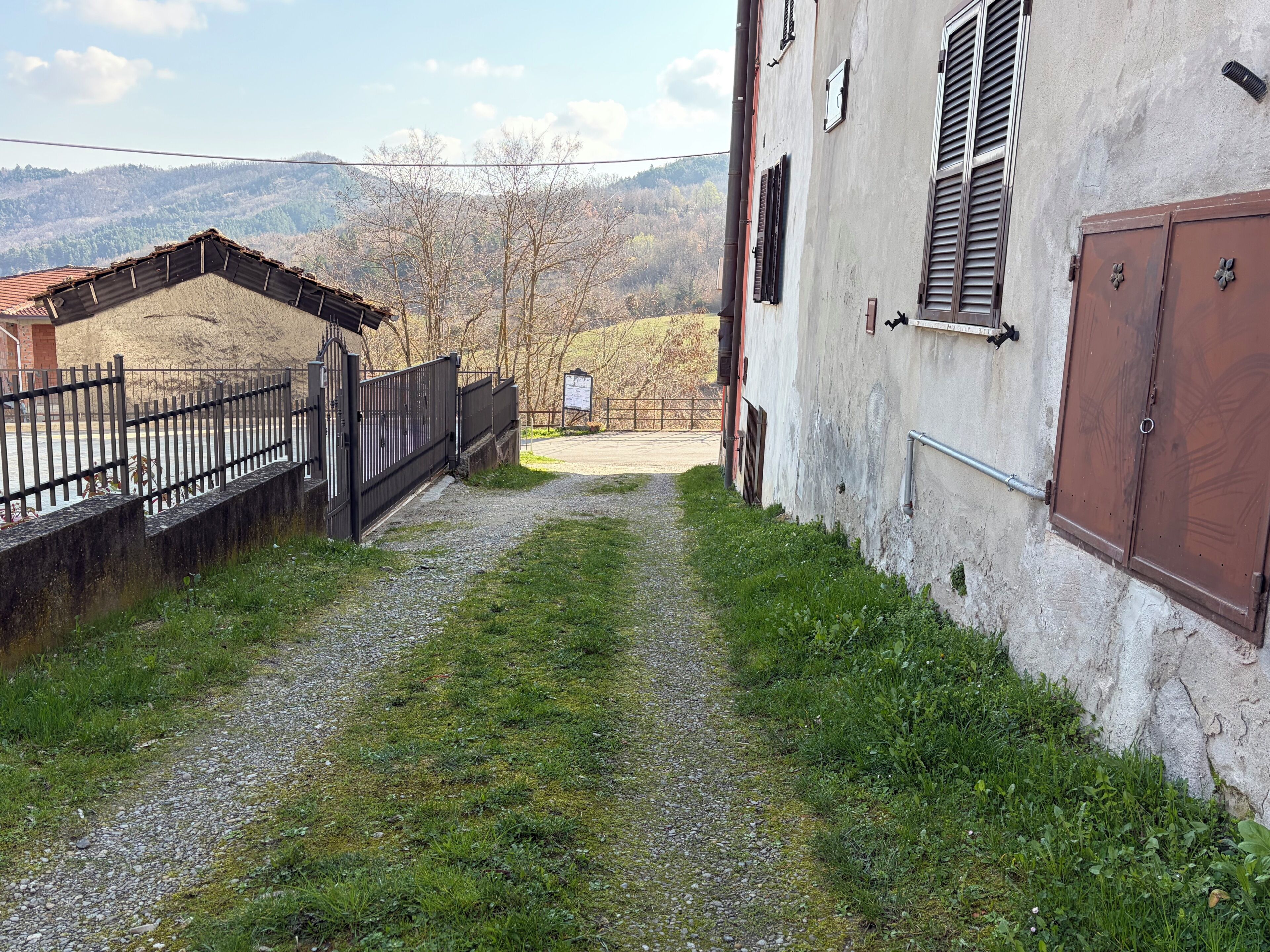 Un étroit sentier de gravier mène à un bâtiment à la façade rustique, doté d’un portail en métal et offrant une vue sur des collines lointaines.