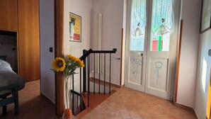 Un couloir avec une porte en bois, un radiateur, un escalier avec une rampe noire et un vase rempli de tournesols.