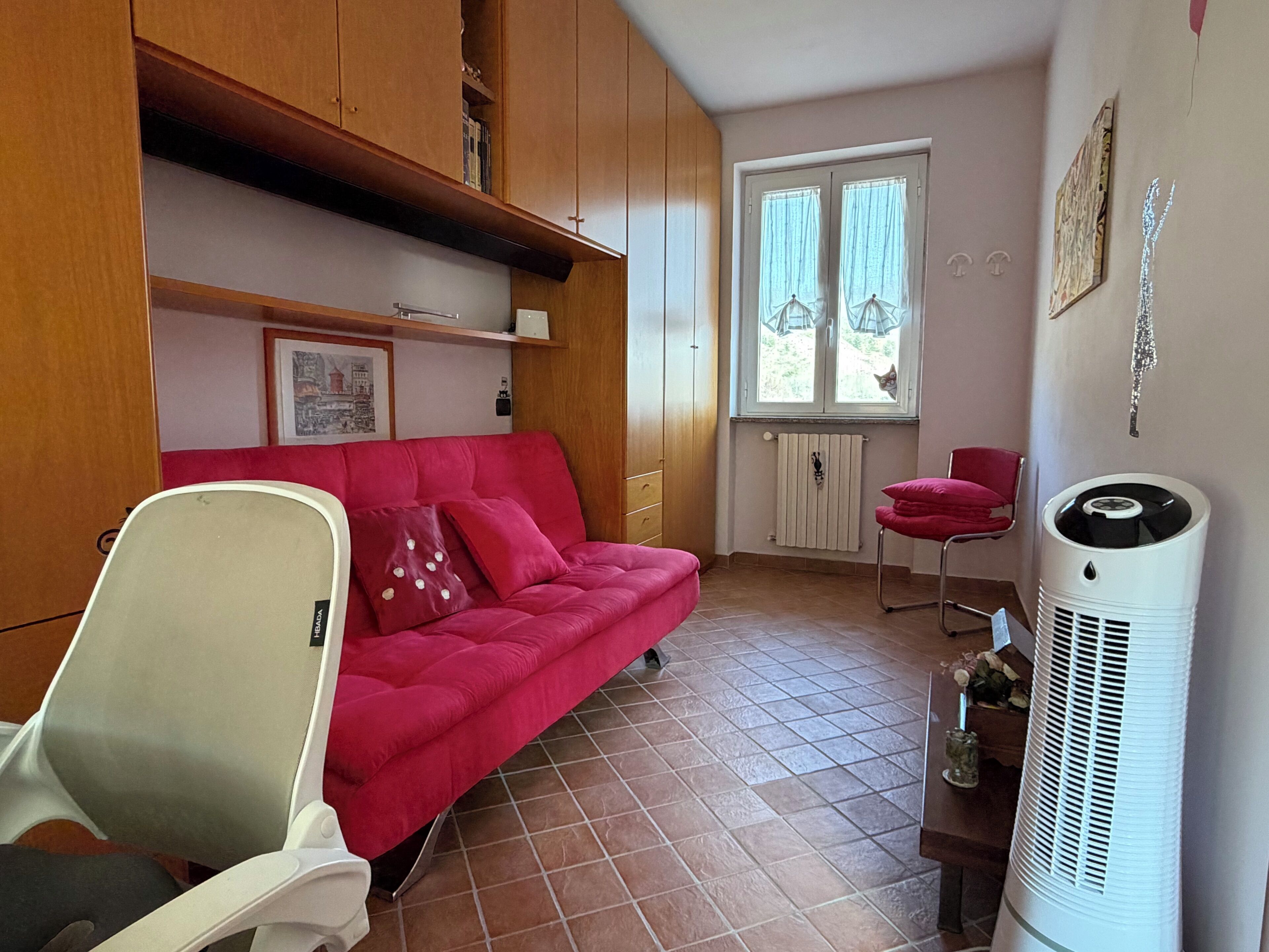 A compact living space with a red sofa, a white chair, a radiator, a window with curtains, and a white air purifier.