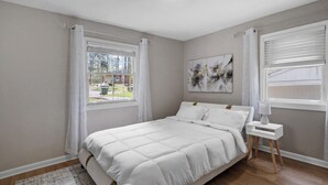 Ein Schlafzimmer mit Bett, Nachttisch, einem Fenster mit Vorhängen und einem Bild an der Wand.