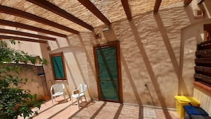 Un patio techado con piso de baldosas, dos sillas blancas de plástico, una puerta y un cubo de basura.