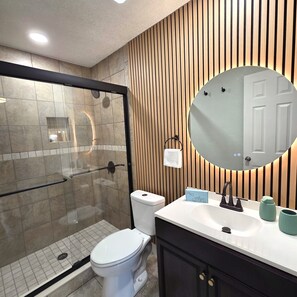 A modern bathroom with a glass-enclosed shower, a toilet, a sink with a mirror, and wooden paneling on the wall. - Serene 1-bedroom apartment in Tampa (Tampa)