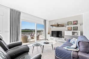 Una sala de estar moderna con sofá, mesa de centro y televisor. Hay un balcón con vistas a las montañas.