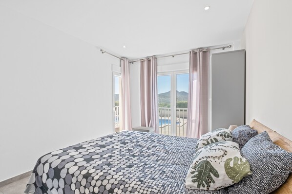 A bedroom with a bed, pillows, a window with curtains, and a view of a pool and mountains.