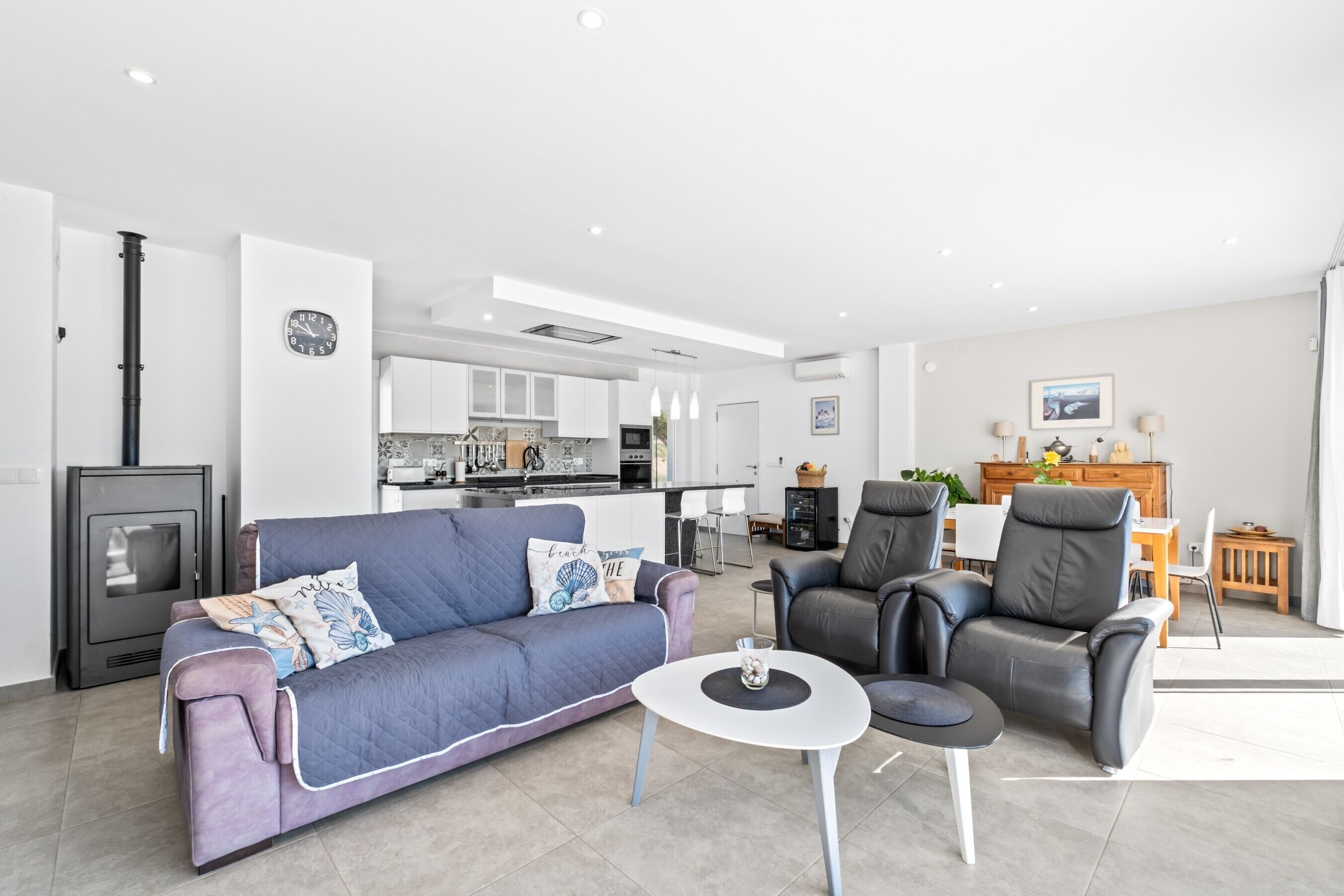 A modern living room with a sofa, chairs, a small table, a fireplace, and a kitchen in the background.
