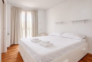 Un dormitorio con cama, cortinas blancas, una ventana con persianas y suelo de madera.