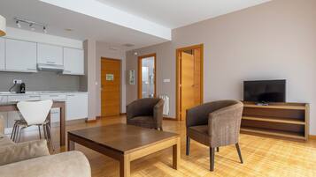 Una sala de estar moderna con una mesa de madera, dos sillones, un sofá, un televisor y una zona de cocina con armarios y una encimera.