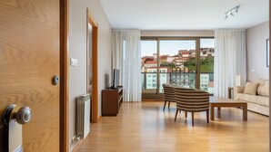Ein modernes Hotelzimmer mit Holzboden, einer Couch, einem gestreiften Sessel, einem Fernseher und Blick auf Gebäude durch große Fenster.