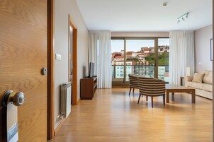 Habitación de hotel moderna con piso de madera, un sofá, una silla a rayas, un televisor y vistas de edificios a través de amplios ventanales.