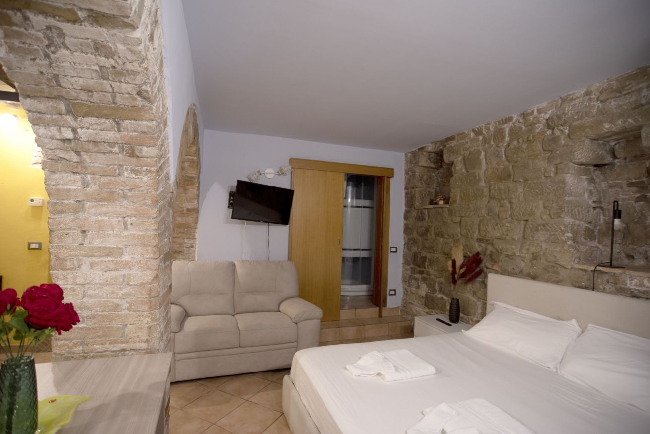 Una habitación con pared de piedra, una cama con ropa blanca, un sofá color beige y un televisor fijado a la pared.