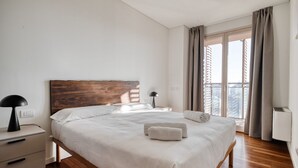 Een slaapkamer met een houten hoofdbord, een bed met witte lakens, een nachtkastje met een lamp en uitzicht door een groot raam.