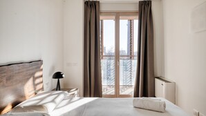 Hotelkamer met een bed, nachtkastje, lamp, radiator en uitzicht op een stadsgezicht door een groot raam.