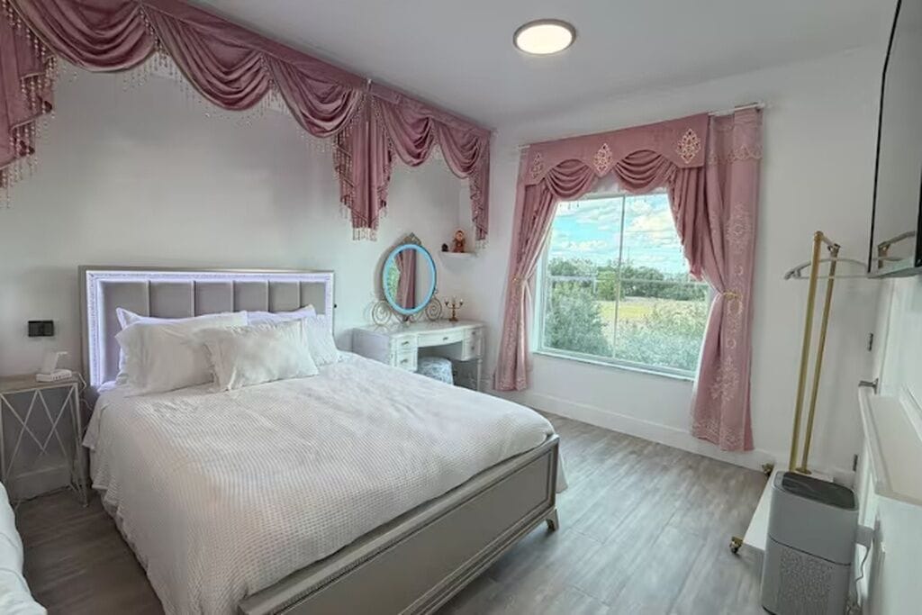 A bedroom with a bed, a vanity, a window with curtains, and a view of greenery outside.