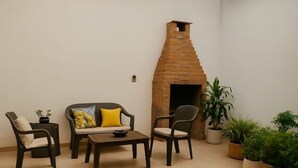 Una habitación con chimenea de ladrillo, un sofá con un cojín amarillo, dos sillones, una mesita de centro y varias plantas en macetas.