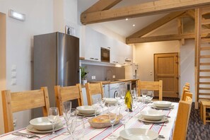A dining area with a table set for six, wooden chairs, a large refrigerator, and a kitchen in the background. - Chalet in Paradiski With Spa & Valley Views (Peisey-Nancroix)