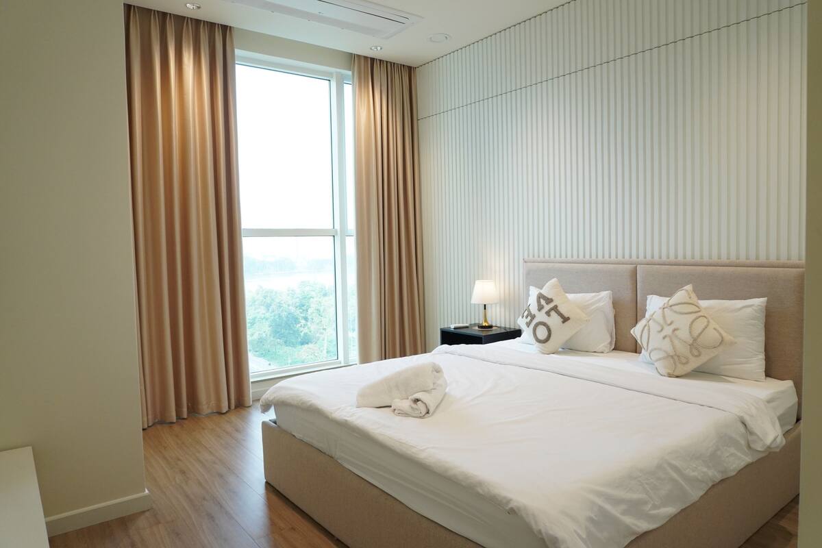 Ein Hotelzimmer mit einem großen Bett, einem Nachttisch mit Lampe und Blick ins Freie durch ein Fenster.