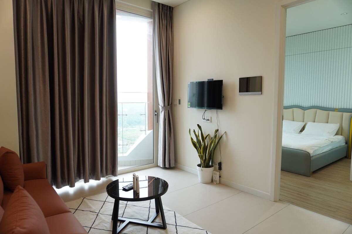 Ein modernes Hotelzimmer mit einer Couch, einem kleinen Tisch, einem Fernseher, einem Bett und einem Balkon mit Vorhängen.