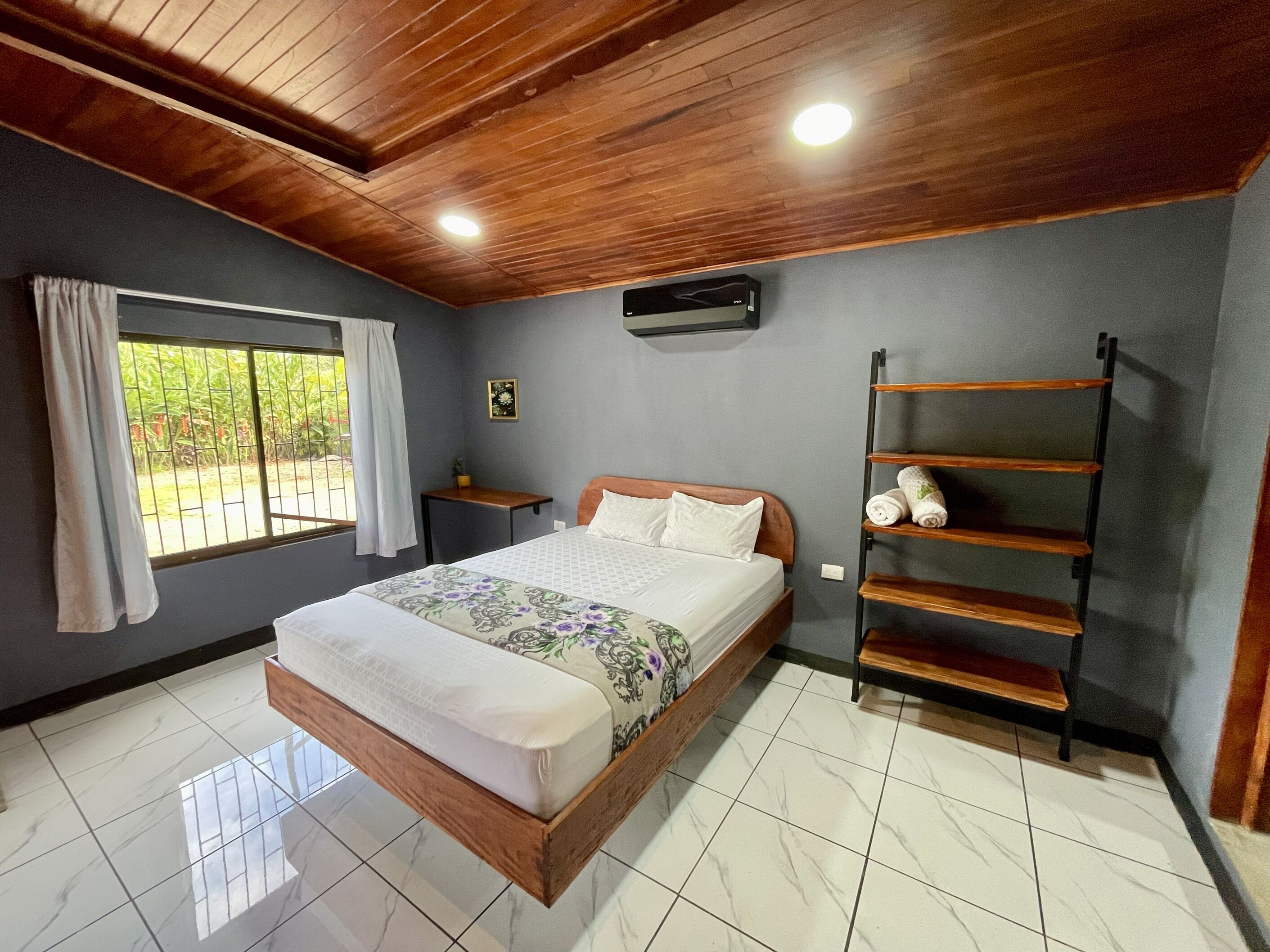 A bedroom with a bed, a window with curtains, a wall-mounted air conditioner, and a wooden shelf.