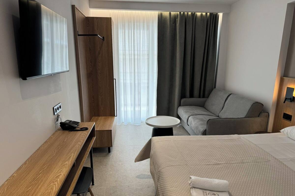 Habitación de hotel con cama, escritorio, sofá, televisión y ventana con cortinas.