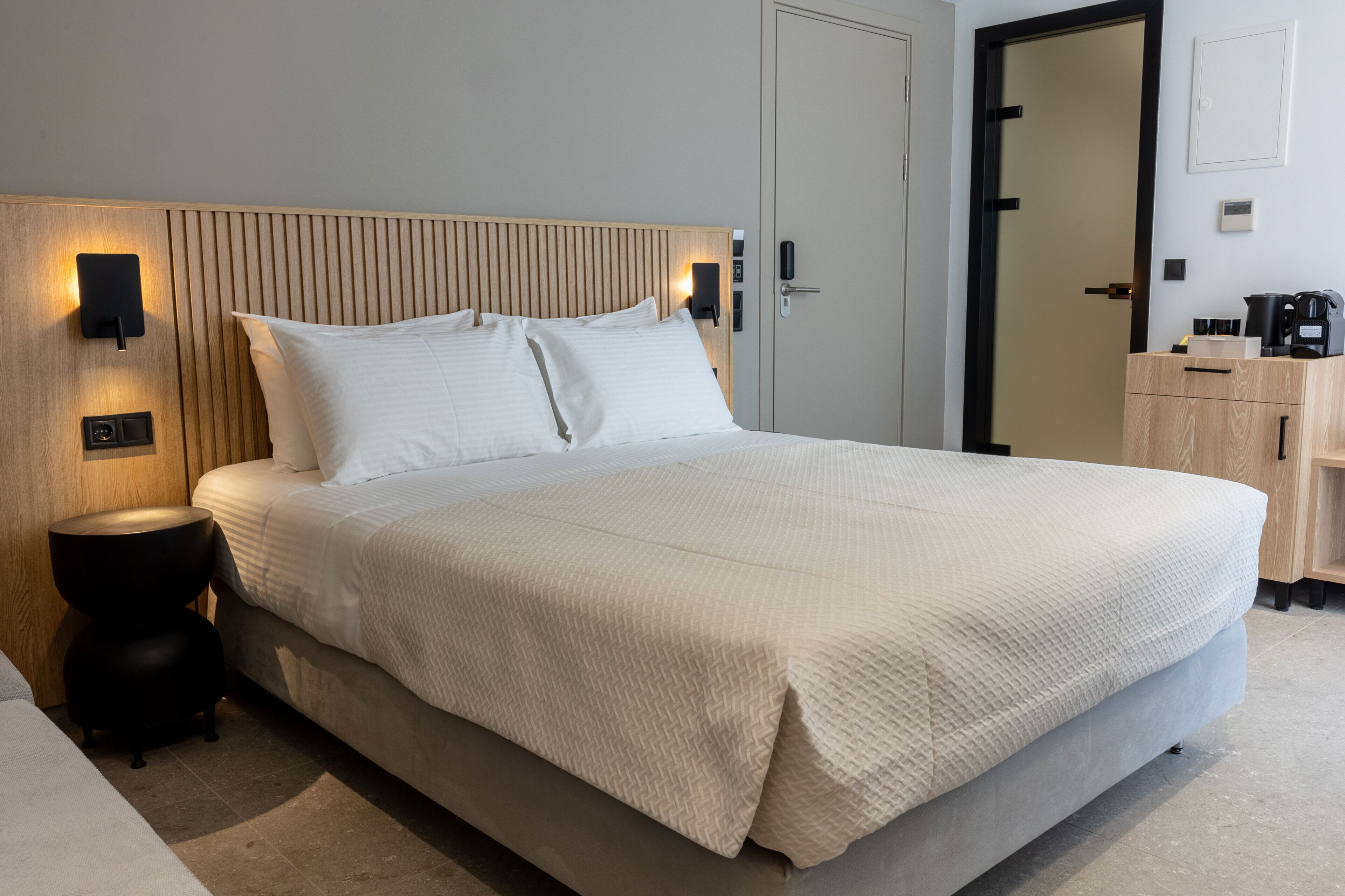 Una cama bien hecha con cabecera, lámparas de noche, una mesita redonda y un aparador de madera.
