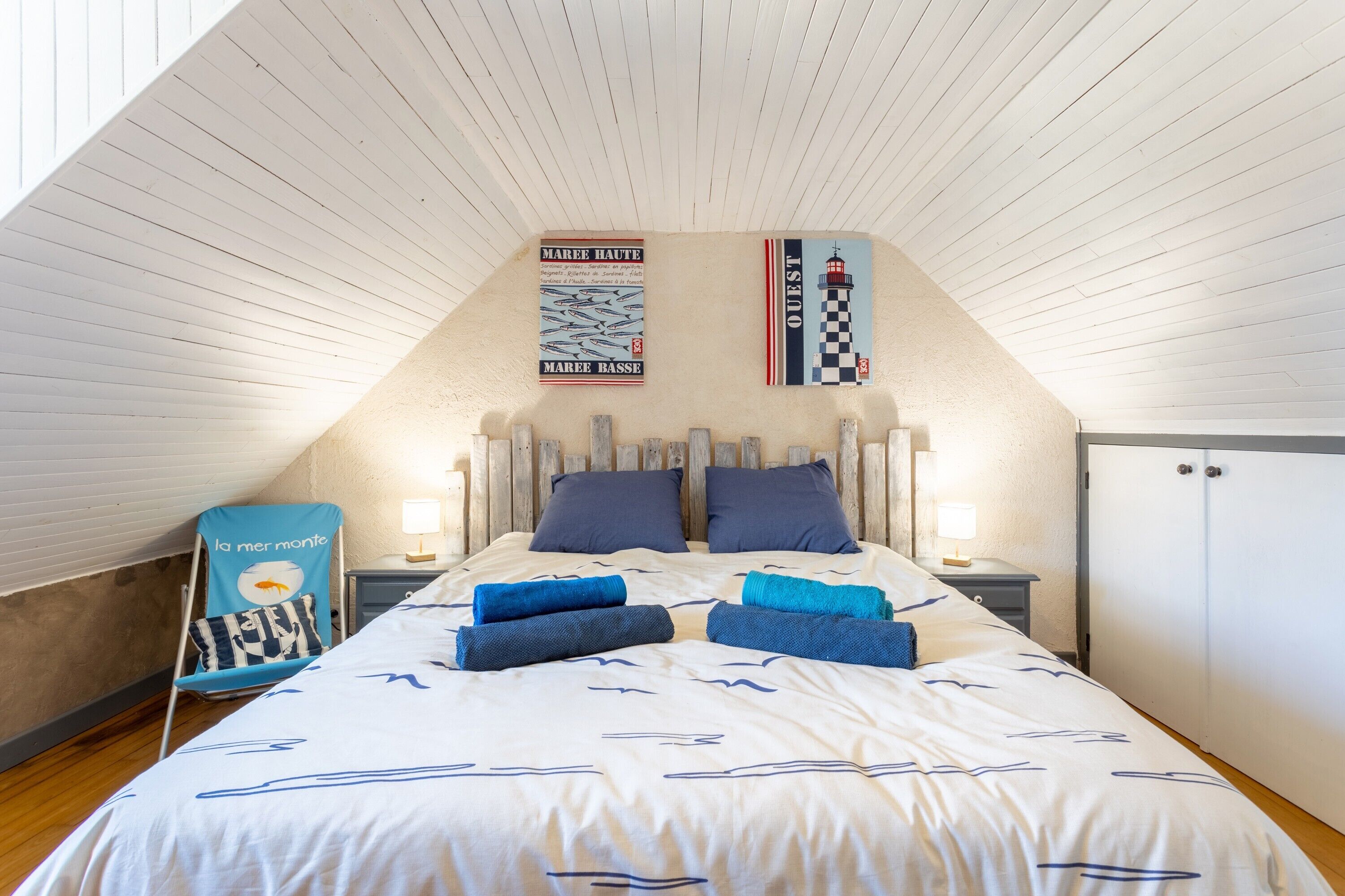 A bedroom with a sloped ceiling, a bed with a nautical-themed duvet, two nightstands with lamps, a wardrobe, and a chair with a fish design.