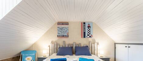 A bedroom with a sloped ceiling, a bed with a nautical-themed duvet, two nightstands with lamps, a wardrobe, and a chair with a fish design.