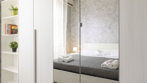 A modern bedroom with a bed, a white wardrobe, and a shelf with books and plants. - Fiera Milano - Total Relax Pero is a cozy and functional apartment, ideal for those seeking tranquility and comfort.Located just minutes from Fiera Milano Rho and well connected to the center of Milan, it offers well-kept rooms, equipped kitchen and comf (Pero)