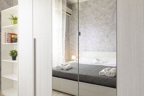 A modern bedroom with a bed, a white wardrobe, and a shelf with books and plants.