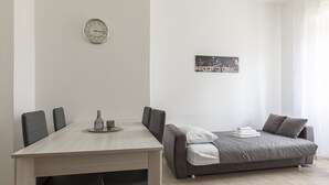 A modern room with a dining table, chairs, a sofa, a bed, a wall clock, and a cityscape picture. - Fiera Milano - Total Relax Pero is a cozy and functional apartment, ideal for those seeking tranquility and comfort.Located just minutes from Fiera Milano Rho and well connected to the center of Milan, it offers well-kept rooms, equipped kitchen and comf (Pero)