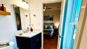 A bathroom with a toilet, sink, and mirror. There is a bed visible through an open door.